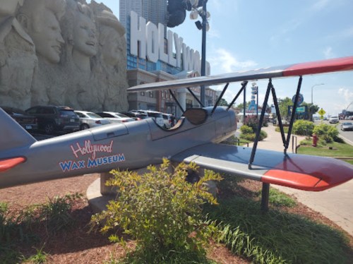 A vintage airplane sculpture outside the Hollywood Wax Museum, surrounded by greenery and city buildings.