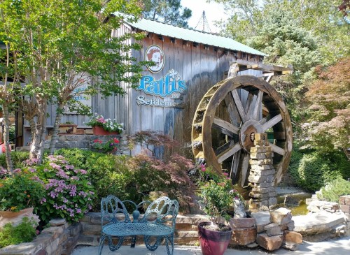 A rustic wooden building with a water wheel, surrounded by colorful flowers and greenery, featuring a sign for "Patti's Settlement."