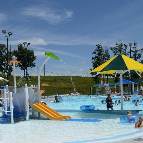 A sunny pool scene with a water slide, splash features, and children playing in the water under colorful umbrellas.