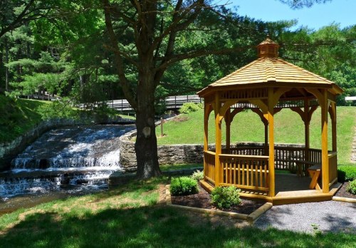 A wooden gazebo beside a flowing stream, surrounded by lush greenery and trees on a sunny day.