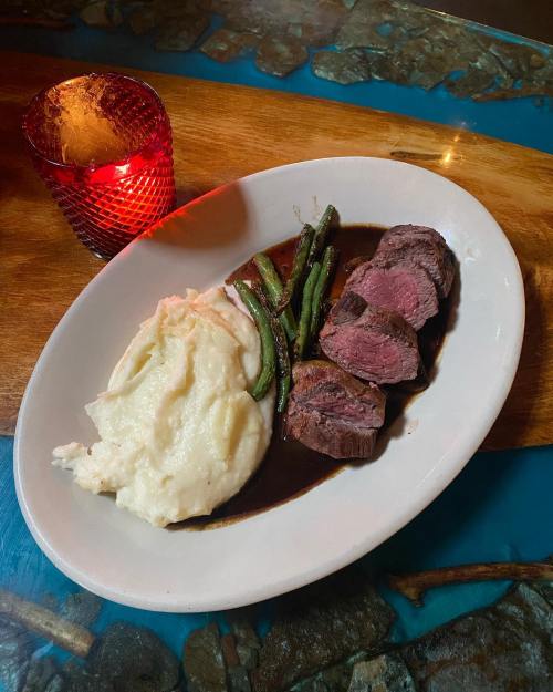 A plate of sliced beef with a rich sauce, green beans, and creamy mashed potatoes, next to a lit red candle.