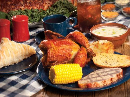 A hearty meal featuring roasted chicken, corn, bread, and soup, served with drinks on a checkered tablecloth.