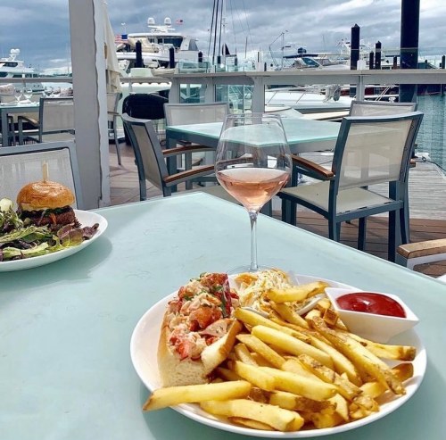 A plate of lobster roll and fries with ketchup, alongside a glass of rosé, set against a marina backdrop.