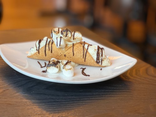 A dessert plate featuring a crispy taco shell filled with cream, topped with toasted marshmallows and drizzled with chocolate.