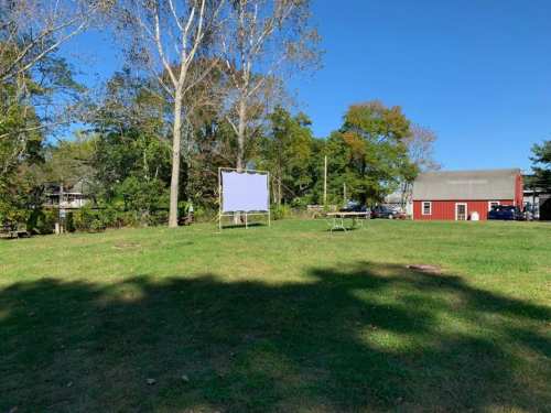 A grassy area with a white screen, picnic tables, and a red barn in the background under a clear blue sky.