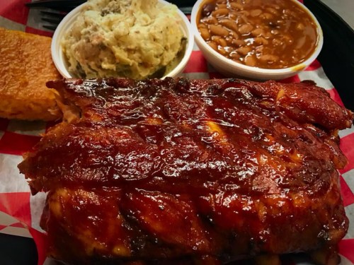 A plate featuring glazed ribs, mashed potatoes, baked beans, and cornbread, all served on a red-checkered tray.