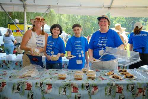 Four people in blue shirts smile behind a table filled with baked goods at an outdoor event.