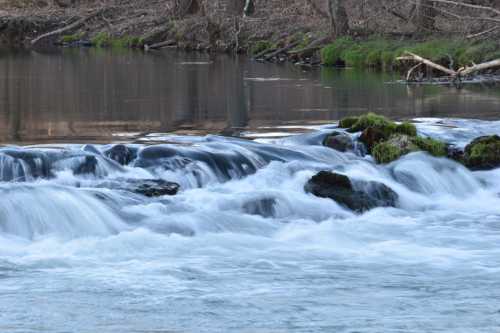 A serene river scene with flowing water over rocks, surrounded by greenery and trees in the background.