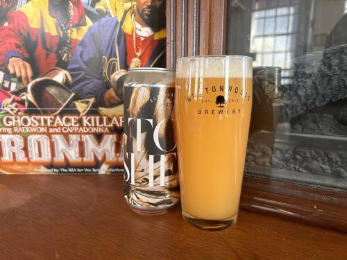 A glass of hazy beer next to a can, with a colorful poster featuring hip-hop artists in the background.
