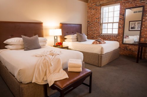 Cozy hotel room with two beds, a mirror, and exposed brick walls, featuring soft linens and decorative pillows.