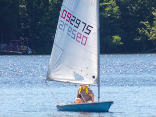 A person sailing on a small boat with a white sail on a calm lake surrounded by trees.
