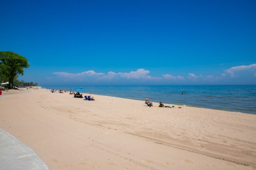 A sunny beach scene with people relaxing on the sand and calm water under a clear blue sky.
