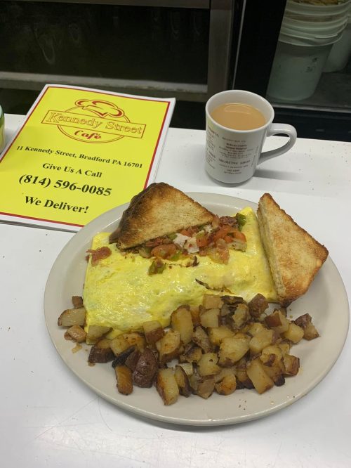 A plate with a large omelet, diced potatoes, and two slices of toast, alongside a cup of coffee and a menu sign.