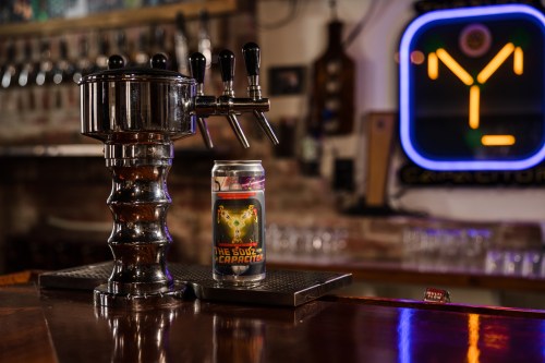 A shiny beer tap with a colorful can of beer on the bar, featuring a neon sign in the background.