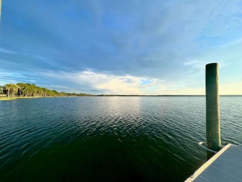 A serene view of a calm lake under a blue sky, with a wooden pier extending into the water.