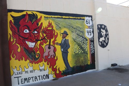 A vibrant mural featuring a devilish figure and a man in a suit, with the text "Lead me not into temptation."