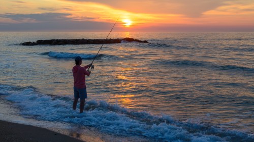 A person fishing at sunset, standing in shallow water with gentle waves and a rocky jetty in the background.