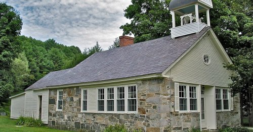 A quaint stone and wood building with a bell tower, surrounded by greenery and under a cloudy sky.