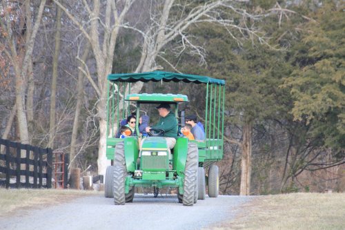 A green tractor pulls a covered wagon with four people along a gravel path in a rural setting. Trees line the background.