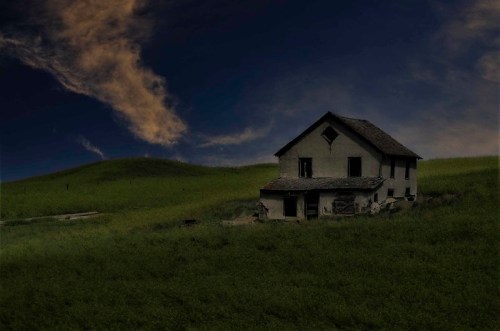 An old, abandoned house sits on a grassy hill under a dramatic sky with clouds.