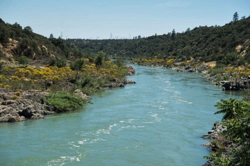 A serene river flows through a lush landscape, surrounded by greenery and rocky banks under a clear blue sky.