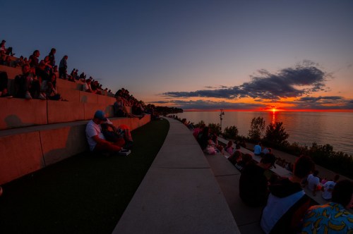 People sitting on a curved concrete seating area by the lake, watching a vibrant sunset over the water.