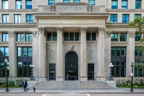 Facade of the John Hancock building featuring grand columns and entrance steps, with two people walking by.