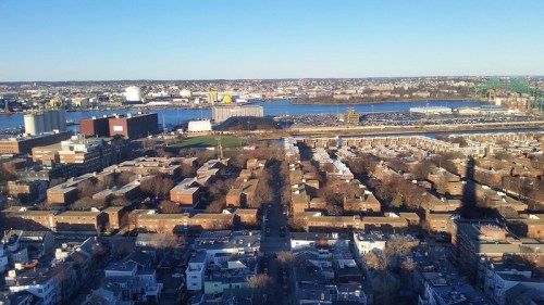 Aerial view of a cityscape with buildings, a river, and industrial areas under a clear blue sky.