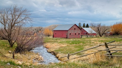 A red barn beside a stream, surrounded by grassy fields and bare trees under a cloudy sky.