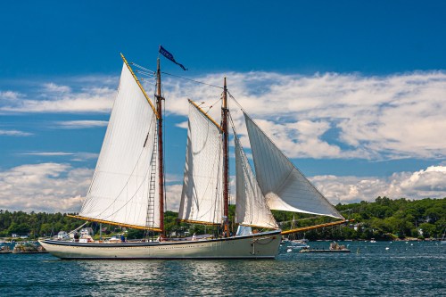 A tall ship with white sails glides through calm waters under a blue sky with fluffy clouds.