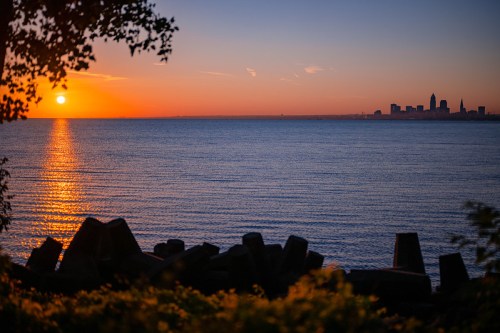 Sunset over a calm lake, with a city skyline in the distance and silhouettes of rocks in the foreground.