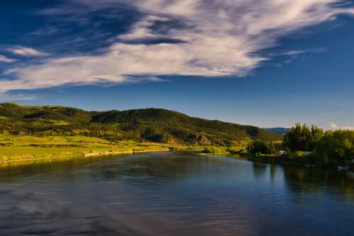 A serene river flows through lush green hills under a blue sky with scattered clouds.