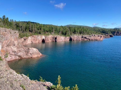 A serene coastal view featuring rocky cliffs, clear blue water, and lush green trees under a bright blue sky.
