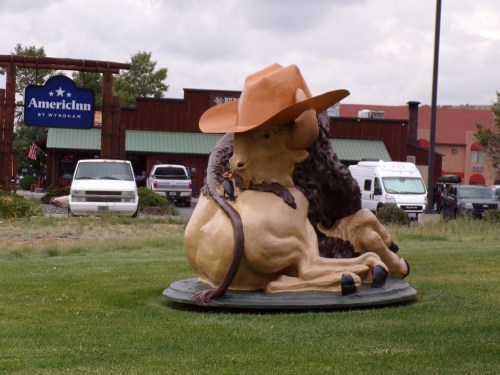 A large statue of a bison wearing a cowboy hat, sitting on a grassy area near an AmericInn hotel.