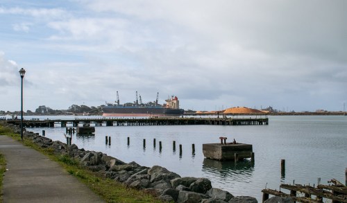 A calm harbor scene with a ship docked, piles of cargo, and a walkway along the water under a cloudy sky.