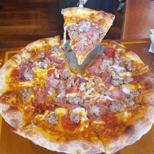 A freshly baked pizza topped with sausage, ham, pepperoni, and melted cheese, with a slice being lifted.