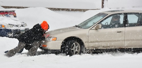 A person in an orange hat pushes a car stuck in deep snow during a snowstorm.