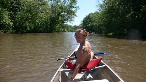 A person paddles a canoe on a calm river surrounded by lush greenery, enjoying a sunny day outdoors.
