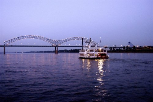 A riverboat glides on water beneath a lit bridge at dusk, with city lights reflecting on the surface.