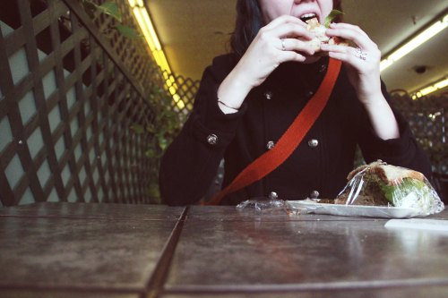 A person in a black coat bites into a sandwich while sitting at a table, with greenery in the background.