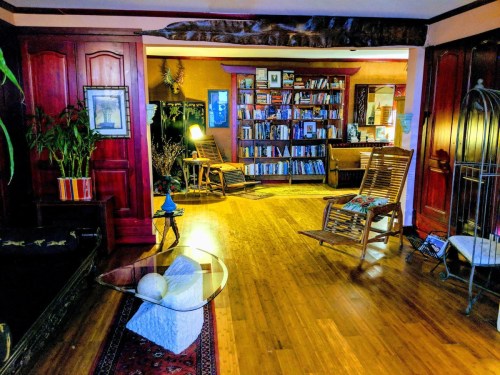 Cozy living room with wooden floors, bookshelves, rocking chairs, and plants, creating a warm, inviting atmosphere.