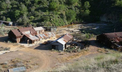 Abandoned buildings with rusted roofs in a green, hilly landscape, near a dry riverbed.
