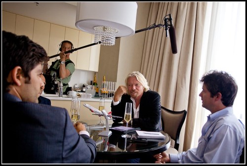 A group of men in a meeting, with one filming and another recording audio, drinks and notes on the table.