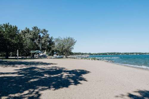 A sandy beach with trees, a pavilion, and a calm lake under a clear blue sky.