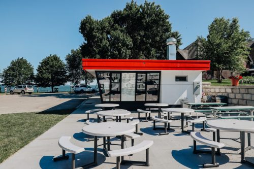 A modern food stand with a red roof, surrounded by picnic tables, near a lake and trees on a sunny day.