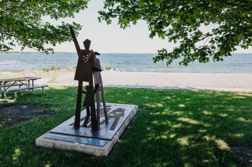 A metal sculpture of two figures stands on a grassy area by the beach, with trees and water in the background.