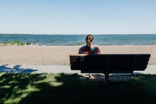 A person sits on a bench by the water, facing the lake with a clear blue sky above and sandy shore in view.