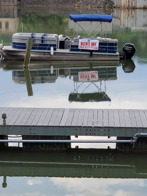 A boat with a "Rent Me" sign is docked at a pier, reflecting in calm water.