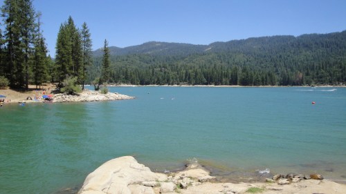 A serene lake surrounded by green trees and mountains, with people enjoying the beach on a sunny day.