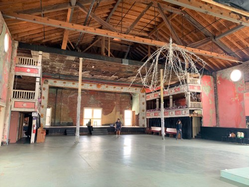 A spacious, rustic interior of a hall with wooden beams, faded walls, and a chandelier made of lights.